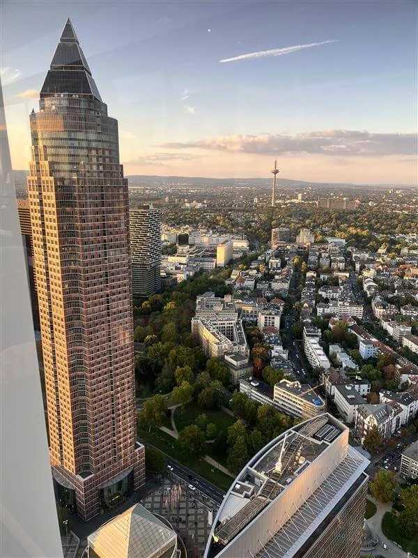 Aerial view of Frankfurt showcasing a prominent skyscraper with a triangular roof design, surrounded by a sprawling urban cityscape. The scene captures a mix of modern architecture and green spaces under a clear sky during daylight. The Frankfurt TV Tower is visible in the background, adding to the city's iconic skyline.