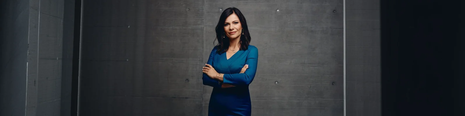 Sladjena Krizaj steht in einem figurbetonten blauen Kleid mit verschränkten Armen vor einer dunklen Betonwand. Die Szenerie erinnert an ein Studio oder einen modernen Industrieraum mit minimalistischer Einrichtung. Ihr Gesicht ist verschwommen, was ihre Anonymität unterstreicht und den Fokus auf Körpersprache und Haltung lenkt. Die Komposition wirkt professionell und seriös, mit kühlen Farbtönen und dramatischer Seitenbeleuchtung.