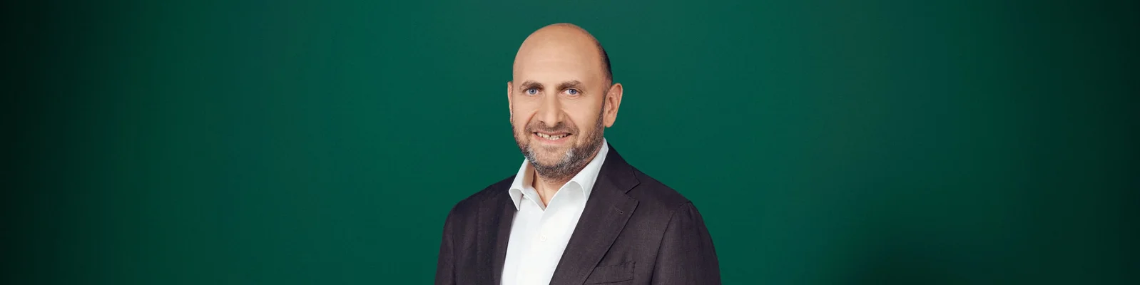 Portrait of a middle-aged man wearing a dark suit and white shirt, standing against a solid green background. He is bald with a trimmed beard and is looking at the camera with a friendly smile. The image has a clean, professional studio look with simple, even lighting. The composition is wide, leaving ample negative space around the subject.