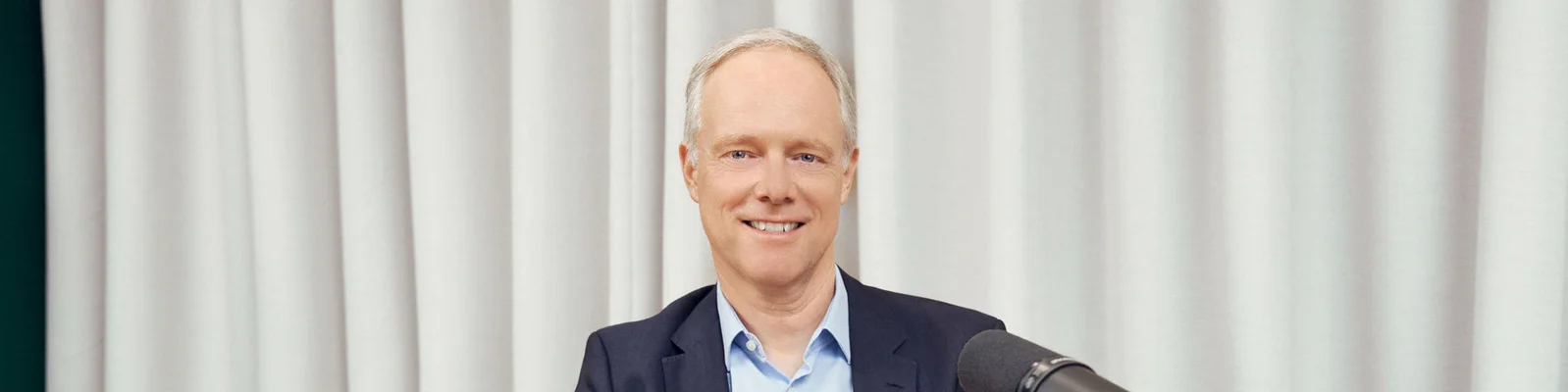 A middle-aged man in a suit jacket sits in front of a microphone, smiling warmly at the camera. He is positioned against a backdrop of light-colored curtains, creating a clean and neutral setting. The composition suggests a podcast, interview, or professional recording environment. The overall mood is approachable, confident, and businesslike.