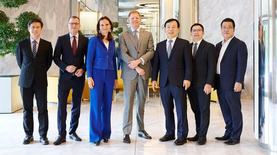 A group of seven individuals dressed in formal business attire stands in a modern, well-lit interior space. The setting features marble walls, large mirrors, and decorative plants, suggesting a corporate or upscale environment. The individuals are positioned in a professional pose, emphasizing a business or networking context.