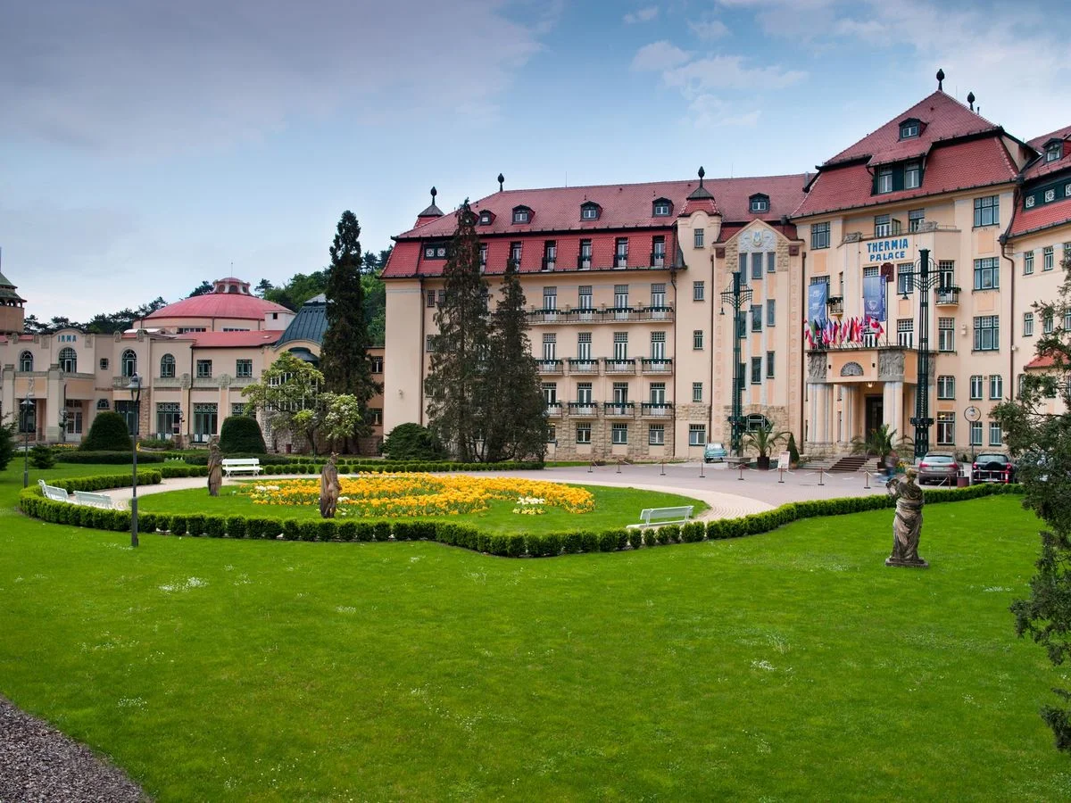 Großes historisches Hotelgebäude mit rotem Dach, umgeben von gepflegten Gärten und Statuen unter wolkigem Himmel.