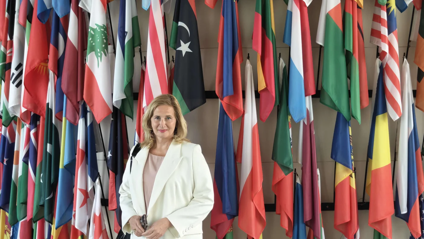 A woman in professional attire stands in front of a dense display of international flags. The scene appears to be in an institutional or conference setting, suggesting a global or diplomatic environment. Brightly colored flags from many countries create a vibrant backdrop behind her. The overall composition emphasizes international cooperation and professional presence.