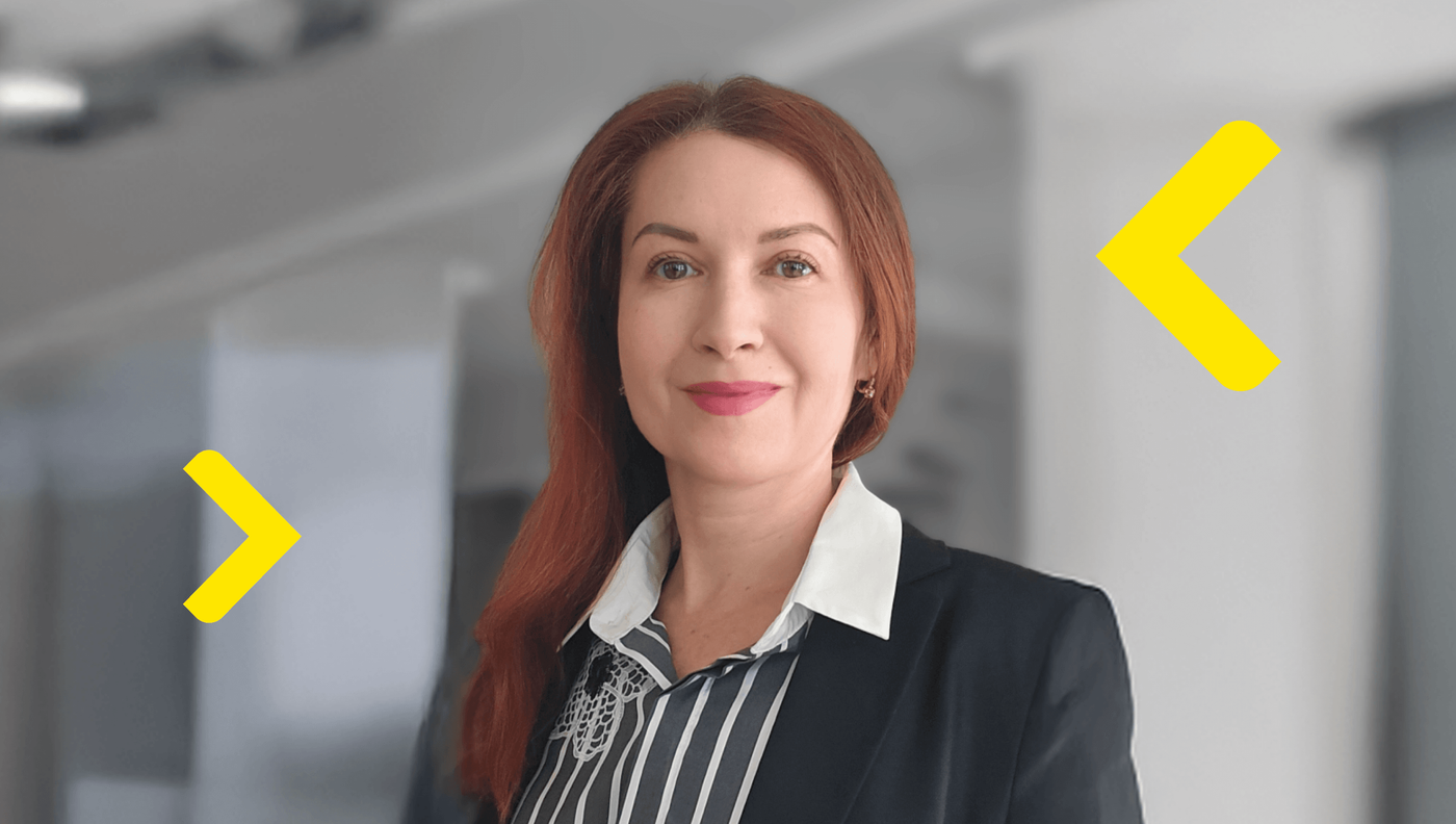 A professional woman with long red hair stands in a modern office corridor, looking directly at the camera with a confident expression. She is dressed in business attire, wearing a dark blazer over a white collared shirt with a patterned top underneath. The background is softly blurred, emphasizing the subject while suggesting a contemporary corporate environment. Bright yellow graphic arrows frame her, adding a dynamic and modern visual element to the composition.