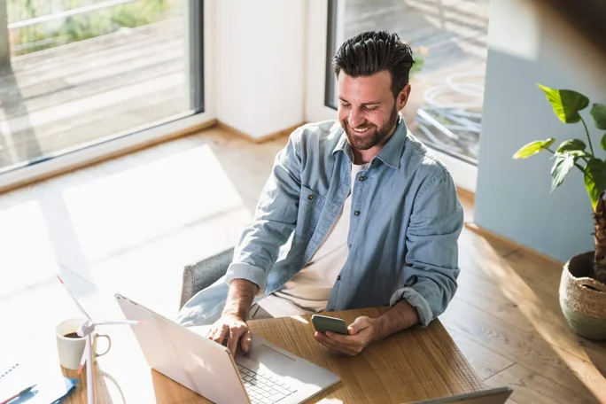 Ein Mann in einem lässigen Jeanshemd sitzt an einem Holztisch und nutzt sowohl einen Laptop als auch ein Smartphone. Sonnenlicht fällt durch große Fenster und erhellt den Raum, in dessen Ecke eine Topfpflanze steht.