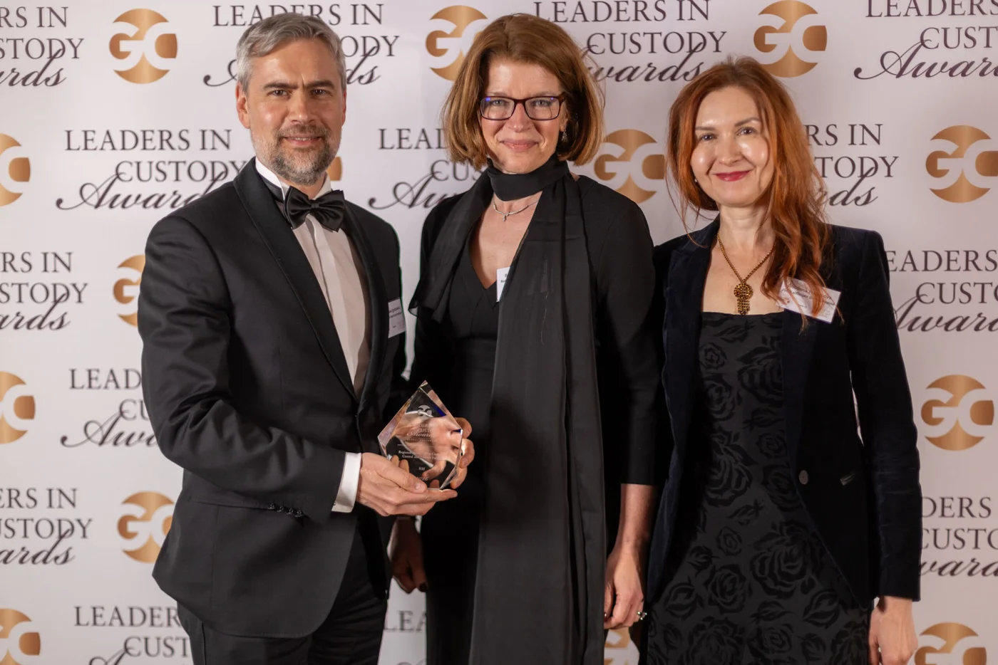 Three formally dressed professionals pose together at an awards event. The central figure stands between two colleagues while one person holds a clear glass trophy. They are positioned in front of a step-and-repeat backdrop featuring the repeated text 'Leaders in Custody Awards.' The scene suggests a corporate recognition ceremony with a celebratory yet professional atmosphere.