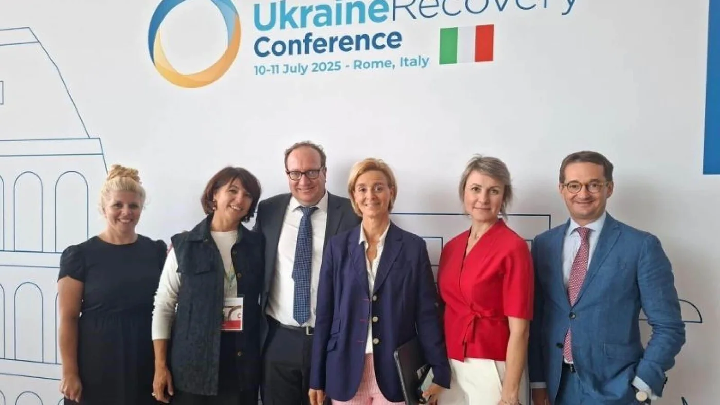 Group of individuals standing in front of a backdrop promoting the Ukraine Recovery Conference. The event is scheduled for July 10–11, 2025, in Rome, Italy, as indicated by visible text. The backdrop features a logo, event name, and Italian flag alongside architectural illustrations.