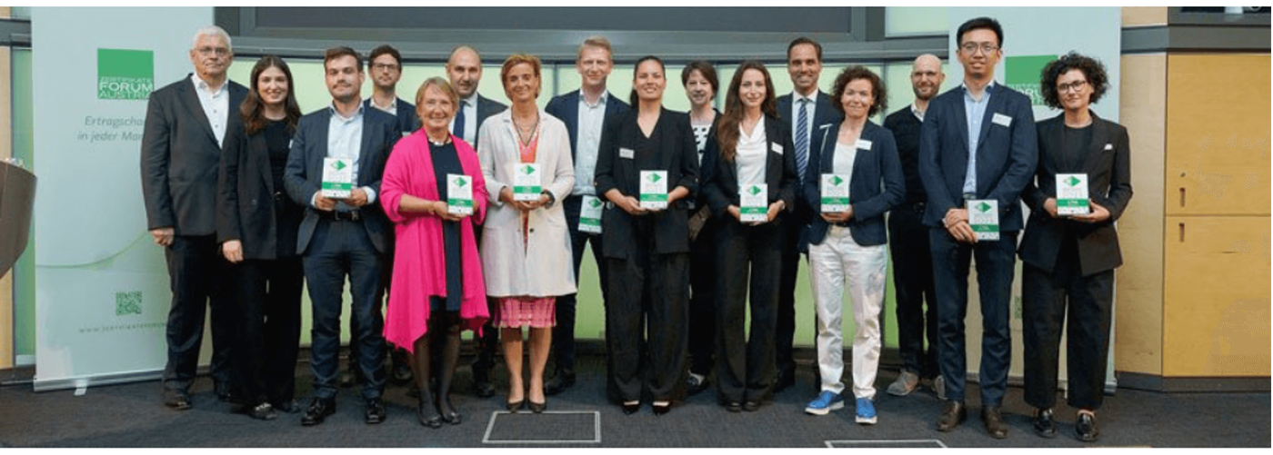 A group of professionals pose together holding awards in a formal event setting. The backdrop features a green banner with visible text 'Empowering people for better lives.' The individuals are dressed in business attire, showcasing a mix of genders and diverse demographics. The awards held by participants display a green and white design.