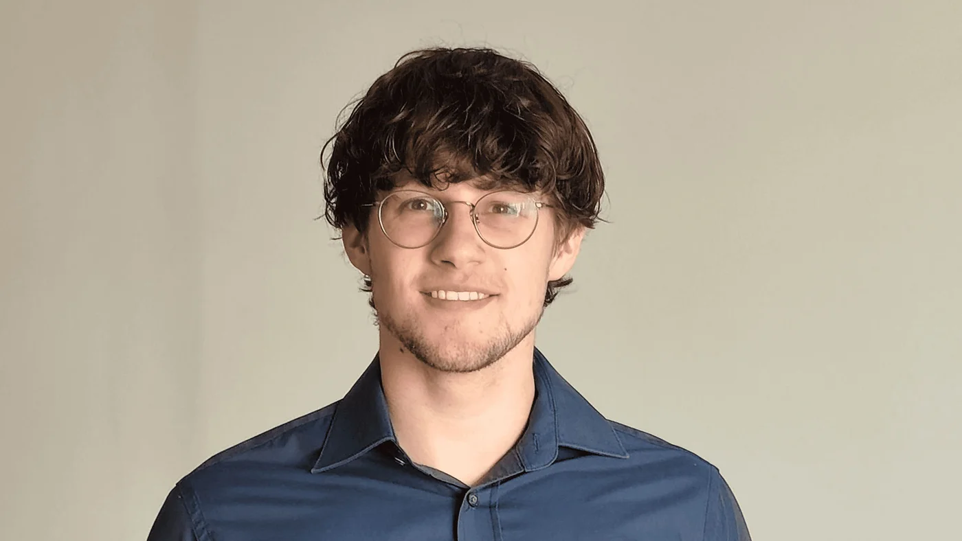 Etienne Van de Moortele wearing a dark blue button-up shirt is standing against a plain, neutral-colored background. The image focuses on the upper body and clothing details. The setting is minimalistic with no additional visible elements.