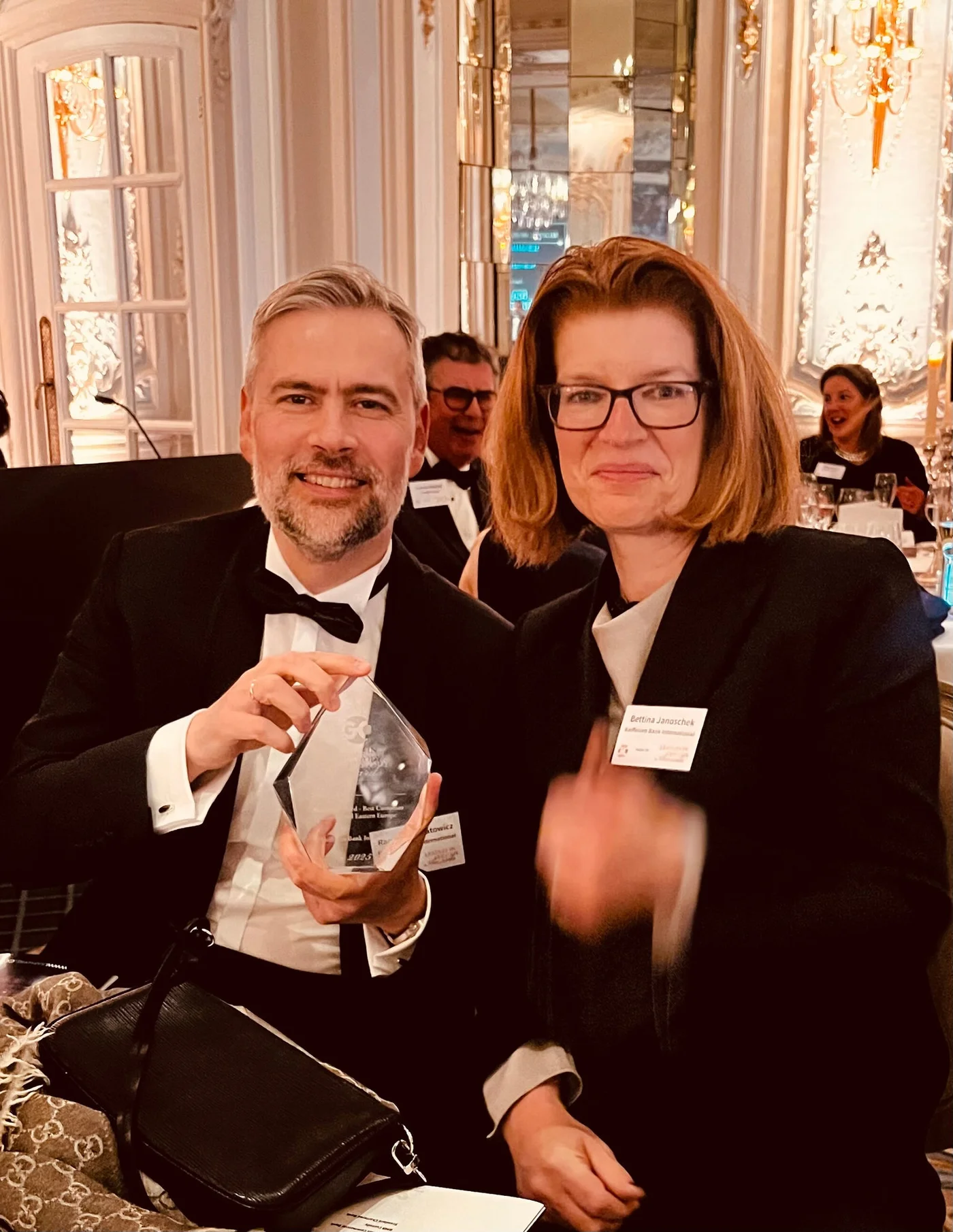 A man and a woman dressed in formal attire sitting at a table in an ornate, elegant setting, with the man holding a glass award.