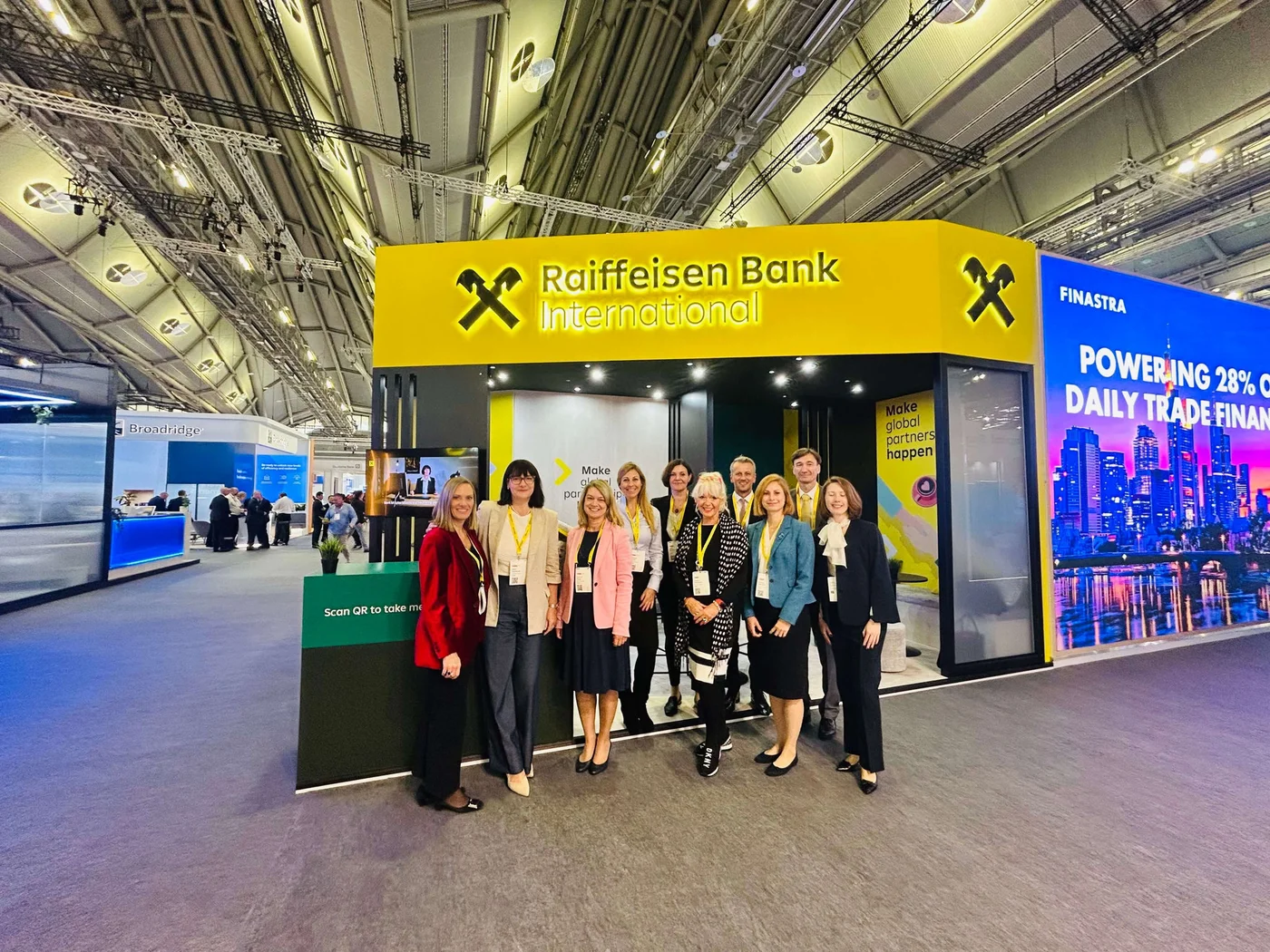 A group of professionals stands in front of the Raiffeisen Bank International booth at a trade event. The booth features bright yellow branding with the company name and logo clearly visible. The setting is an indoor exhibition hall with modern lighting and other booths in the background. Visible text includes 'Raiffeisen Bank International' and 'Powering 28% of daily trade finance'.