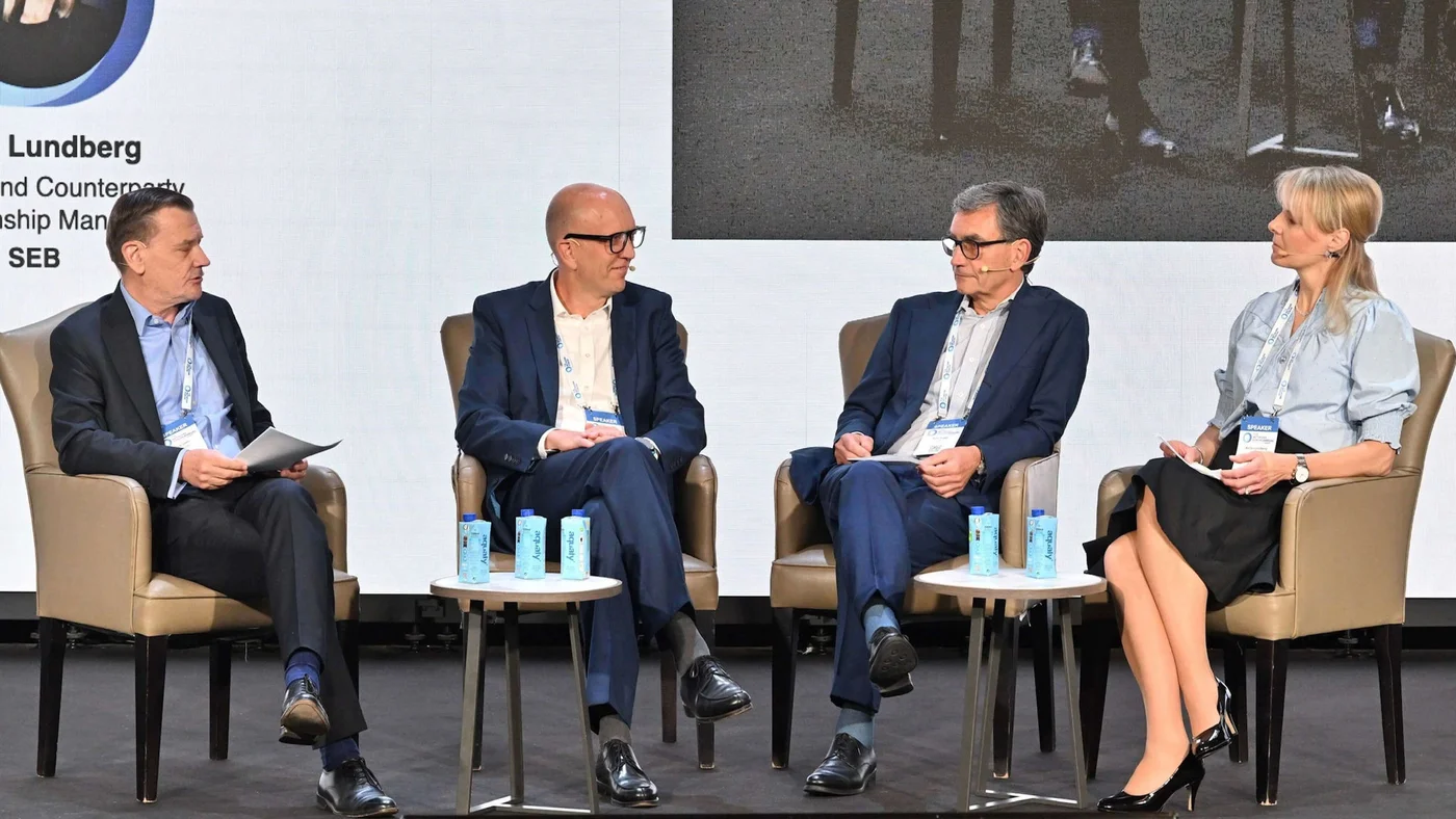 Eine Gruppe von vier Geschäftsleuten sitzt während einer Podiumsdiskussion auf der Bühne. Die Szenerie umfasst Stühle, kleine Tische mit blauen Getränkedosen und eine Projektionsleinwand im Hintergrund. Die Teilnehmenden sind formell gekleidet, und die Umgebung deutet auf ein Unternehmens- oder Konferenzumfeld hin.