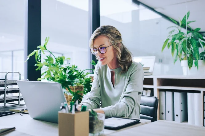  Eine Fachkraft, die an einem Schreibtisch arbeitet, umgeben von Pflanzen, zeigt einen modernen und produktiven Büroraum, der Natur für bessere Konzentration und Wohlbefinden integriert.