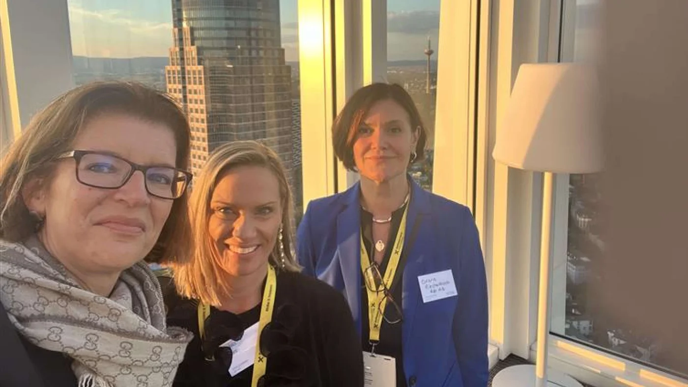 Eine Gruppe von Personen nimmt an einer beruflichen Netzwerkveranstaltung in einem Hochhaus teil. Im Hintergrund erstreckt sich die Skyline mit einem markanten Wolkenkratzer und dem warmen Schein des Sonnenuntergangs. Eine moderne Stehlampe trägt zur eleganten Atmosphäre bei. Gelbe Namensschilder und Lanyards deuten auf ein formelles Firmentreffen hin.