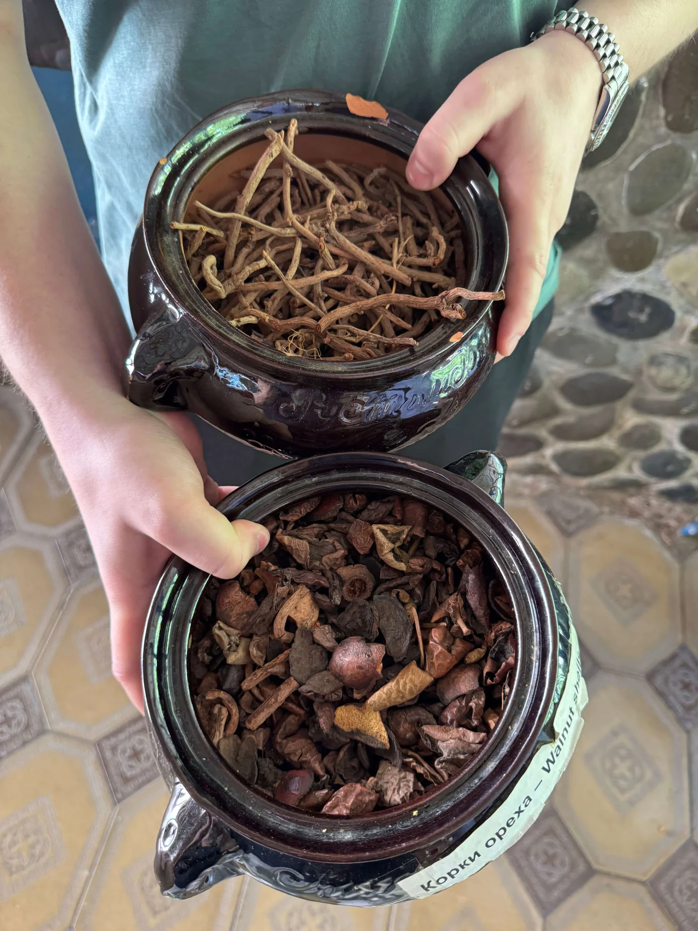 A person holds two dark ceramic jars filled with dried plant materials. The upper jar contains long, thin roots or stems, while the lower jar is filled with mixed dried bark and pieces. The person’s hands and watch are visible against a patterned floor and stone wall background. A small label with Cyrillic text is attached to one of the jars, suggesting an herbal or natural remedy context.