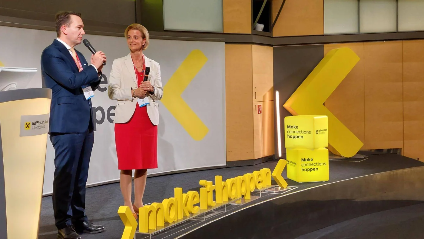 Zwei Personen mit Mikrofonen sprechen auf der Bühne bei einer Firmenveranstaltung mit gelbem "make it happen"-Schriftzug und Raiffeisen Bank-Branding.