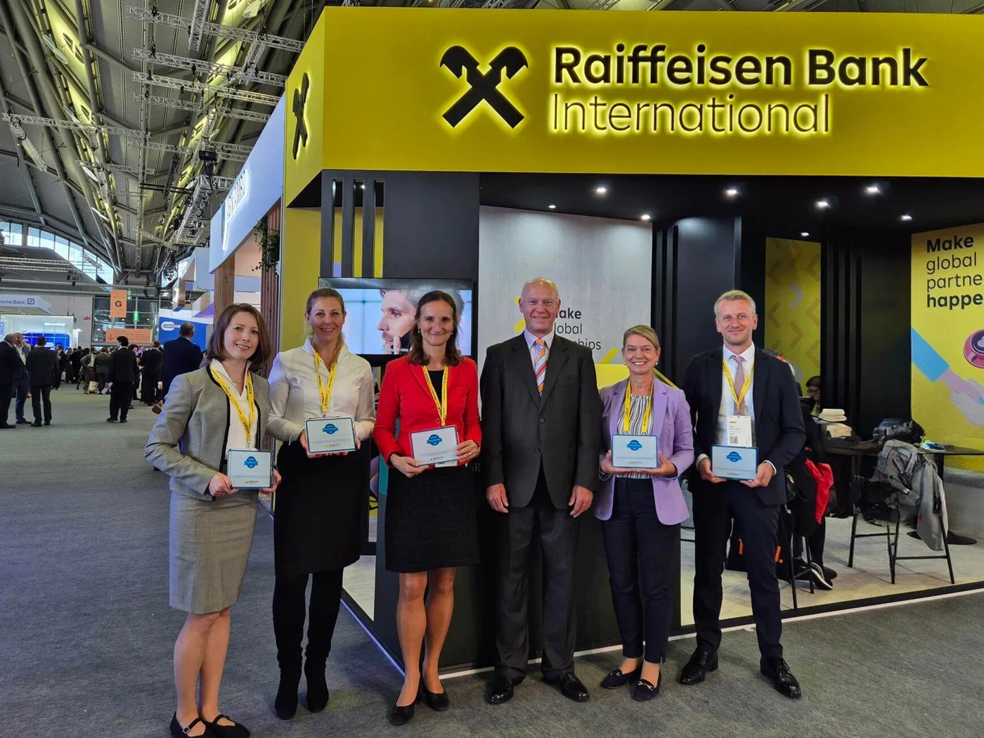 Group of individuals posing in front of the Raiffeisen Bank International booth at a professional event. The booth features bright yellow branding and text, with visible signage and a modern design. Participants are holding plaques, and the setting appears to be a corporate or networking event in a large indoor venue.