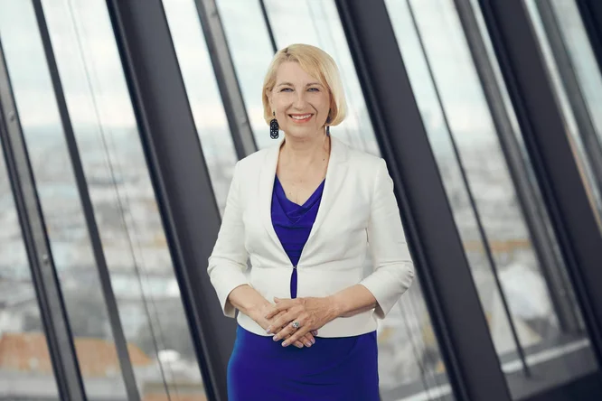 Andrea Gaal steht lächelnd vor bodentiefen Fenstern in einem modernen Bürogebäude. Sie trägt ein hellblaues Kleid mit einem weißen Blazer und auffälligen Ohrringen. Im Hintergrund ist eine leicht verschwommene Stadtkulisse zu sehen, die das Motiv hervorhebt. Das natürliche Licht, das durch die Fenster fällt, schafft eine klare, moderne Büroatmosphäre.