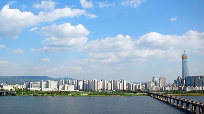 Panoramablick auf eine moderne Stadtlandschaft mit hohen Wolkenkratzern und einer Brücke, die einen breiten Fluss überspannt, unter einem Himmel mit fluffigen Wolken.