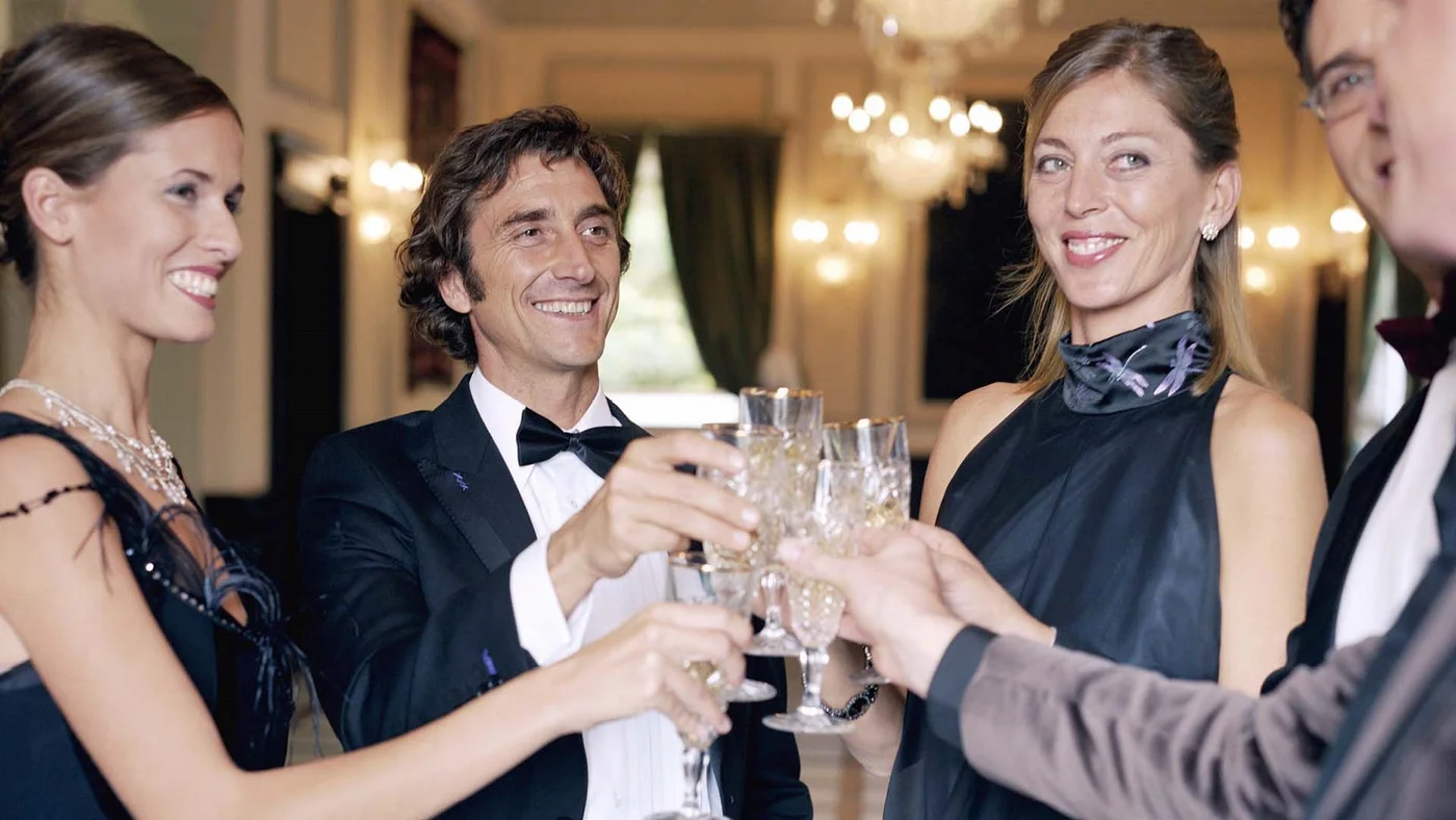Eine Gruppe elegant gekleideter Menschen, die bei einer formellen Veranstaltung mit Sektgläsern anstoßen, vor einem luxuriösen Hintergrund mit Kronleuchter.