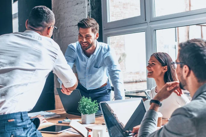 Eine Gruppe von Fachleuten in einem modernen Büro schüttelt sich die Hände und diskutiert Ideen an einem Tisch mit Laptops und Dokumenten, was Teamarbeit, Partnerschaft und Geschäftserfolg repräsentiert.