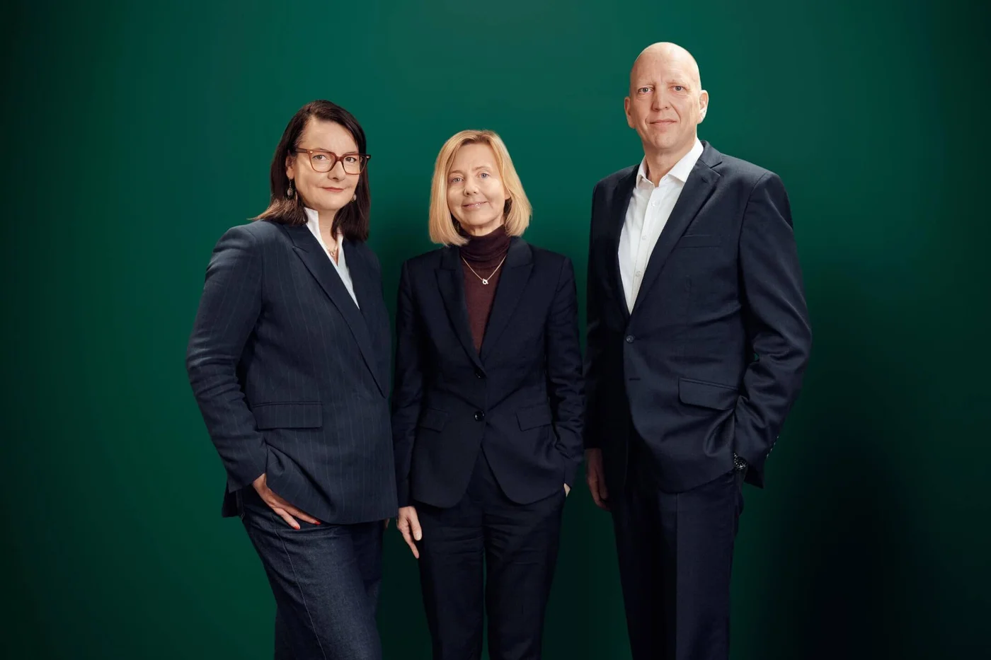 Eine Gruppe von drei Erwachsenen – zwei Frauen und ein Mann – steht vor einem einfarbig grünen Hintergrund. Alle tragen dunkle Anzüge und wirken dadurch formell und professionell. Die Studiobeleuchtung unterstreicht die seriöse Atmosphäre. Es sind keine Texte, Logos oder Zahlen zu sehen.