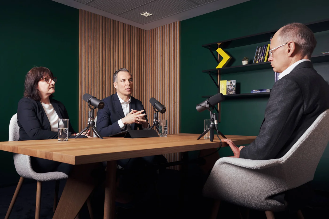 Eine Gruppe von Personen sitzt um einen Holztisch mit Mikrofonen und führt eine Podcast-Diskussion. Die Umgebung hat eine grüne Wand und Holzvertäfelung, was eine professionelle Atmosphäre schafft.