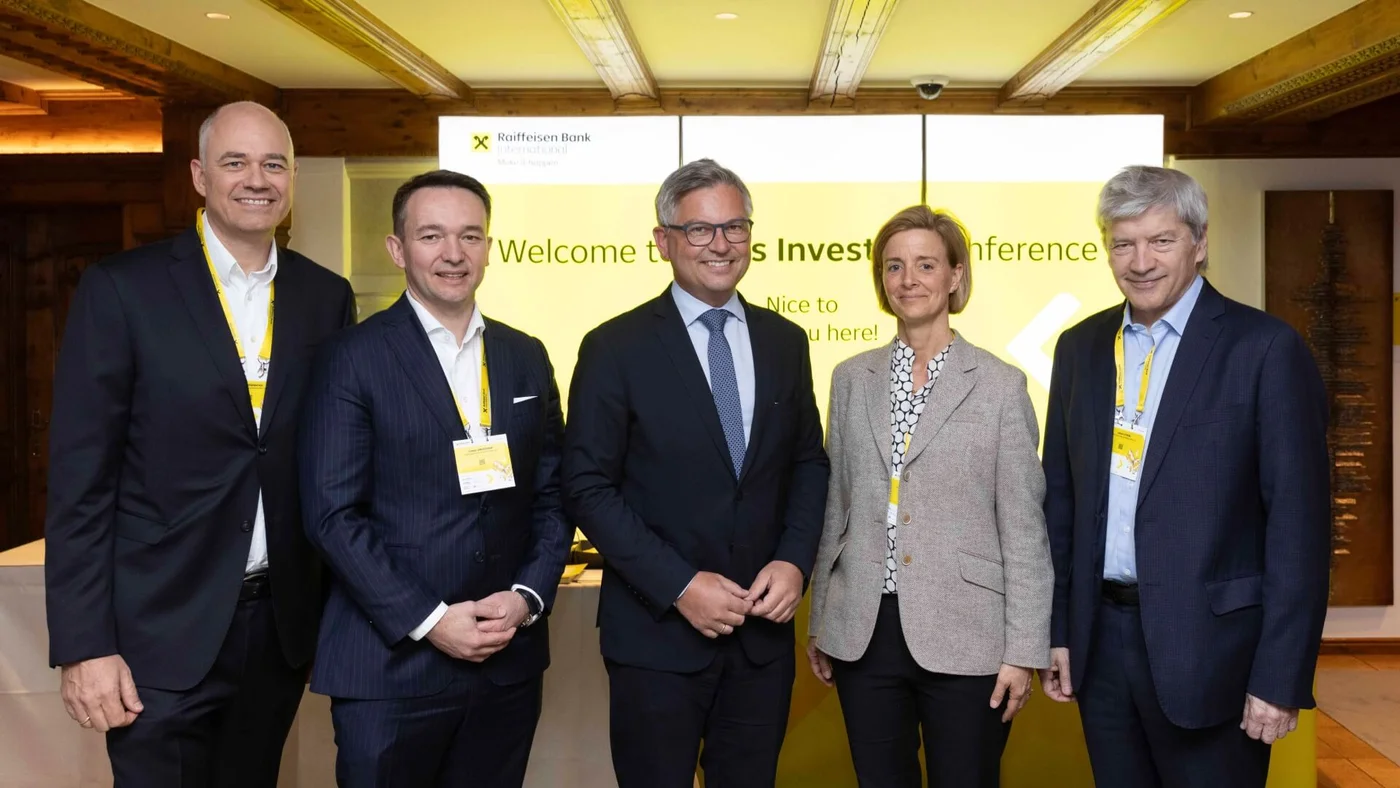 Eine Gruppe von fünf Berufstätigen in Anzügen steht vor einem gelben Hintergrund mit dem Raiffeisen Bank Logo bei einer Fachkonferenz.
