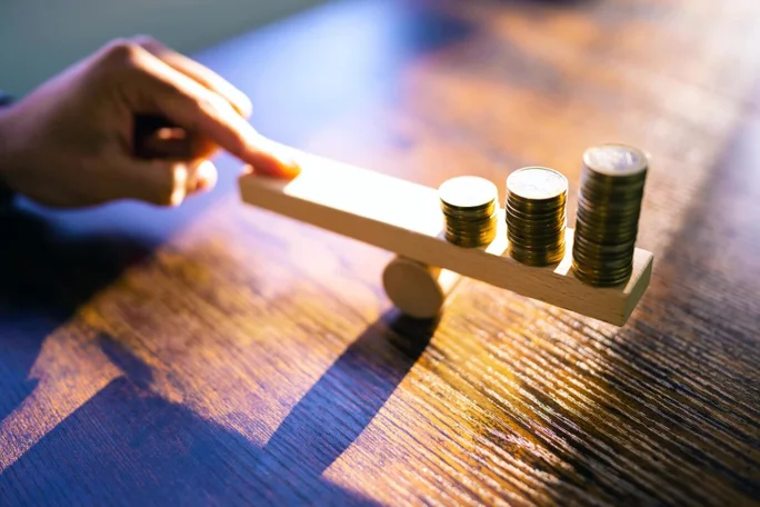 Dieses Bild zeigt eine Hand, die einen hölzernen Hebel balanciert, auf dessen beiden Seiten sich Stapel von Münzen befinden. Dies symbolisiert das Konzept der Abwägung finanzieller Investitionen und die sorgfältige Überlegung, die bei der Verwaltung von Vermögenswerten und Ressourcen erforderlich ist.
