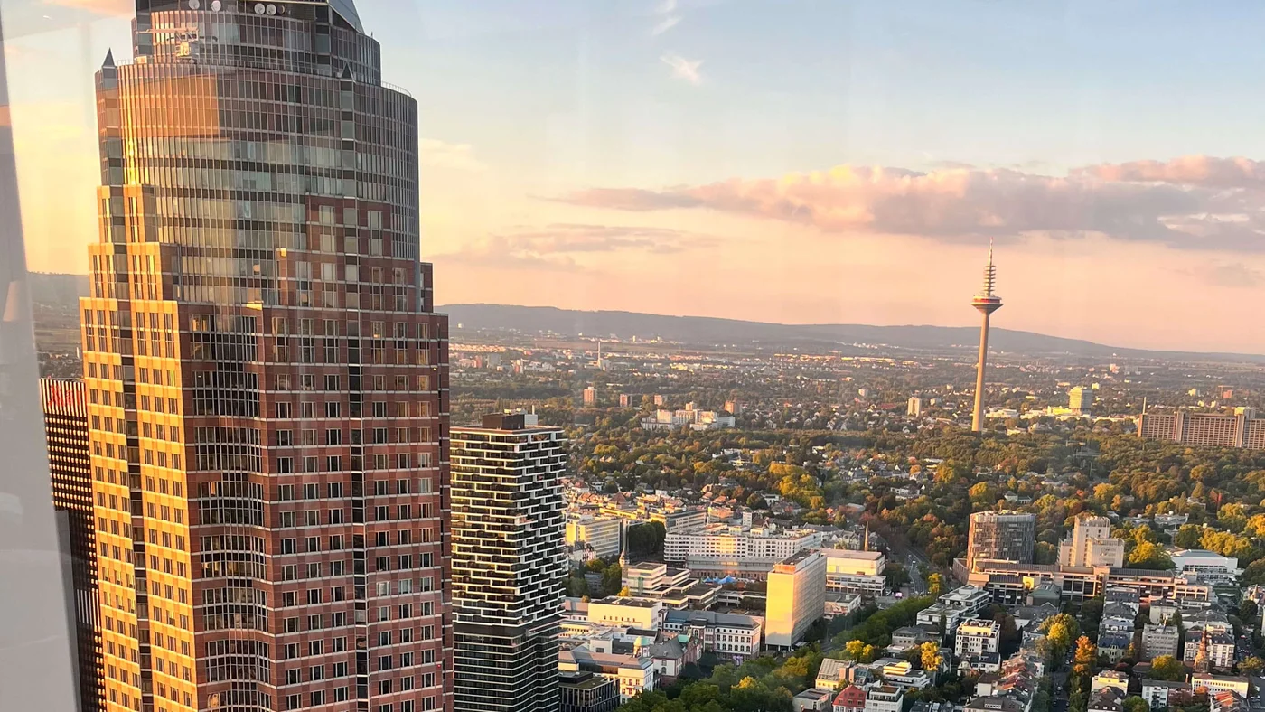 Ein Hochhaus dominiert den Vordergrund und präsentiert moderne Architektur in Frankfurt am Main. Die Szenerie fängt einen warmen Sonnenuntergang mit vereinzelten Wolken ein, die die Stadt in goldenes Licht tauchen. Der ikonische Frankfurter Fernsehturm ist in der Ferne zu sehen, umgeben von üppigem Grün und urbaner Bebauung.