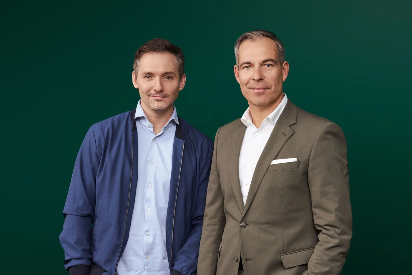 Henry Murray von Waterdrop und Daniel Rath sind formell gekleidet: Er trägt einen blauen Blazer und ein blaues Hemd, er einen braunen Anzug mit weißem Hemd. Der einfarbig grüne Hintergrund erzeugt eine professionelle und klare Bildwirkung. Das Foto unterstreicht den geschäftlichen oder formellen Charakter des Bildes.