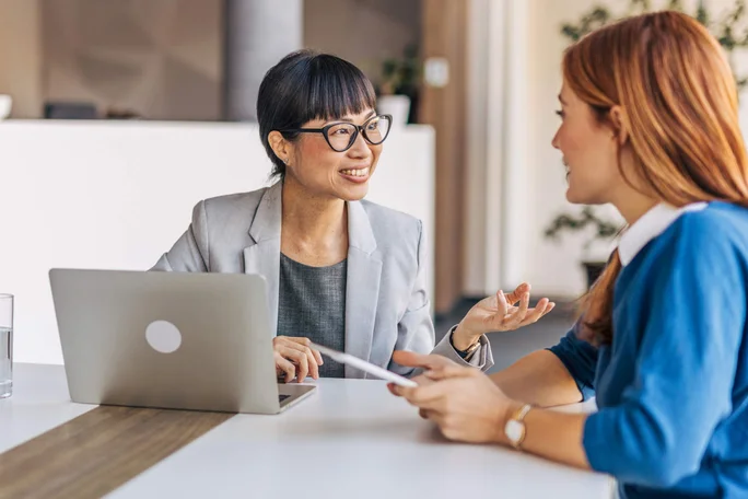 Zwei Frauen führen eine Diskussion an einem modernen Bürotisch. Eine trägt einen grauen Blazer mit einem Laptop vor sich, während die andere ein Tablet hält, was auf eine kollaborative Arbeitsumgebung hinweist.