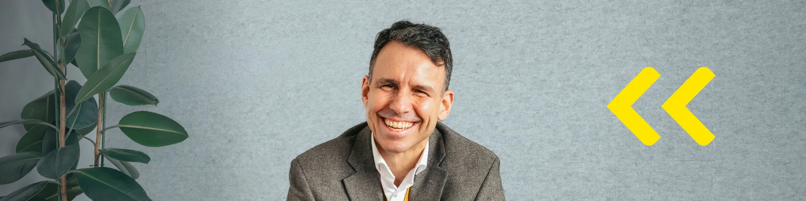 A business professional sits against a light gray wall, partially obscured by a blur over the face. A large green indoor plant is visible on the left side of the frame. On the right, two bold yellow arrows point toward the person, adding a graphic design element. The overall scene suggests a modern office or corporate setting with a clean, minimal background.
