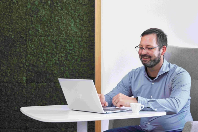 Ein Mann in einem blauen Hemd sitzt an einem weißen Schreibtisch und arbeitet an einem Laptop. Die moderne Einrichtung besticht durch eine grüne, strukturierte Wand und eine kleine weiße Kaffeetasse auf dem Tisch. Die Szene vermittelt eine professionelle und konzentrierte Atmosphäre.