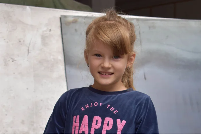 Ein Kind steht draußen und trägt ein marineblaues T-Shirt mit der Aufschrift „Enjoy the Happy“ in rosa Buchstaben.