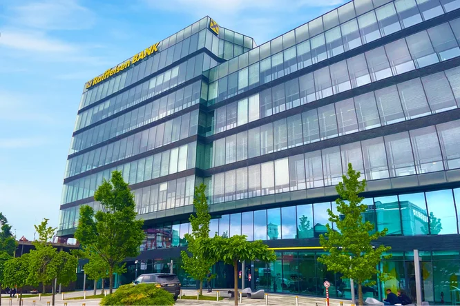 Das schlanke, moderne Design des Bürogebäudes der Raiffeisen Bank Kosovo mit großen Glasfenstern und gepflegter Außenanlage vor klarem, blauem Himmel.