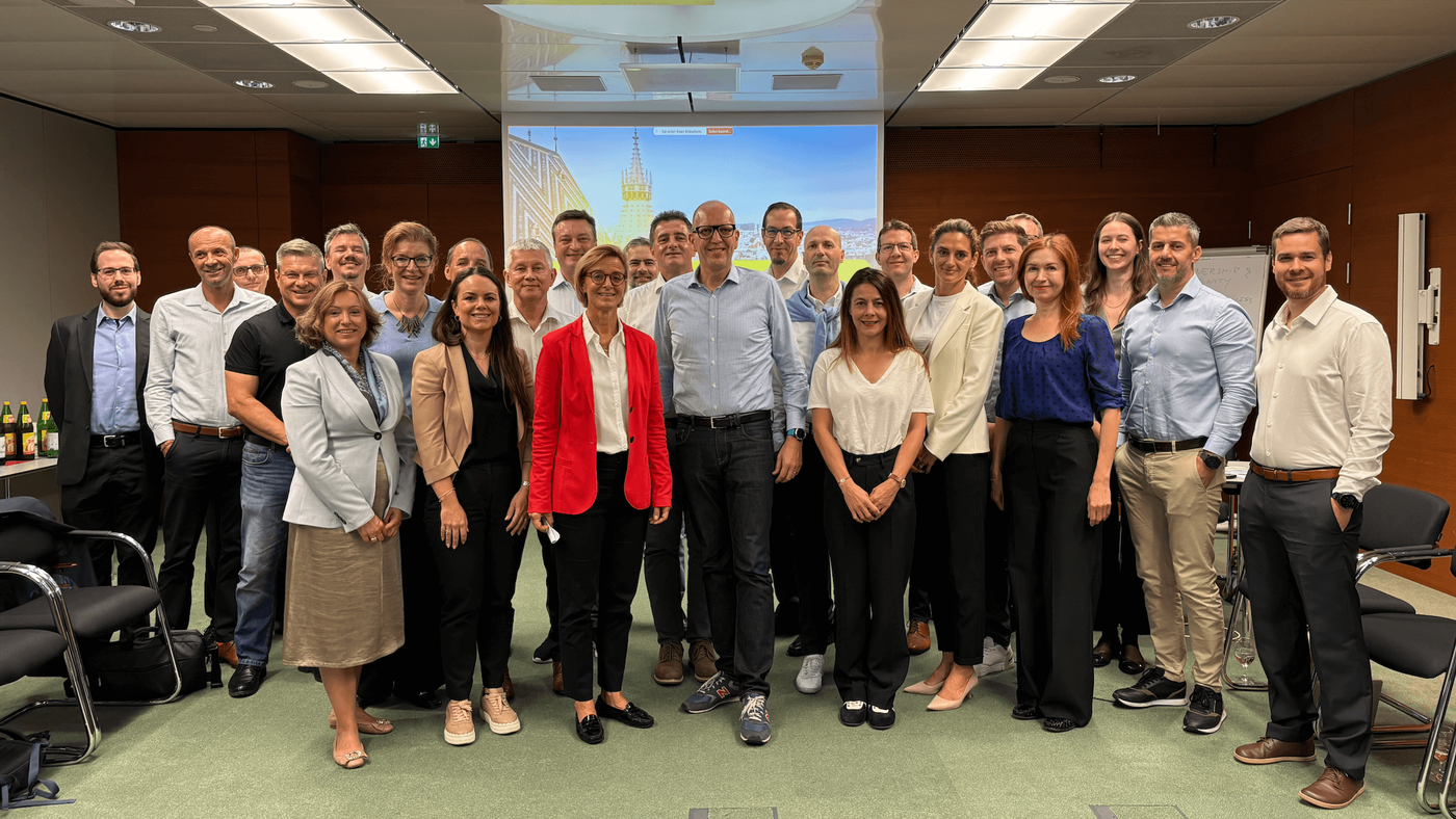 Eine Gruppe von Fachleuten posiert für ein Foto in einem modernen Konferenzraum. Auf der Leinwand ist eine Stadtansicht mit einem Uhrturm zu sehen. Die Teilnehmer tragen Business-Casual-Kleidung. Der Raum ist mit Stühlen und einer holzvertäfelten Wand ausgestattet.