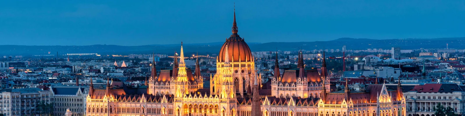 The Hungarian Parliament Building glows brightly against a twilight sky in Budapest, Hungary. The iconic architecture features intricate details and spires, surrounded by the cityscape. The warm lighting contrasts beautifully with the cool blue tones of the evening.