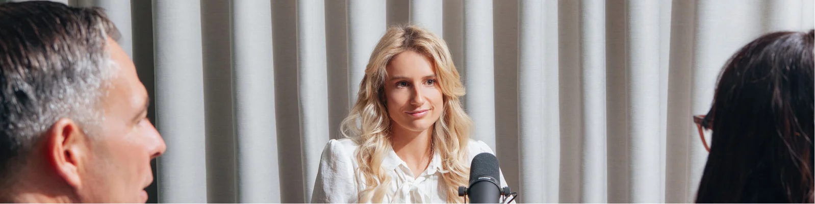 Anna Gasser spricht bei einer formellen Veranstaltung und steht vor einem Mikrofon. Im Hintergrund sind neutrale Vorhänge zu sehen, was auf einen Innenraum hindeutet. Im Vordergrund sind zwei Personen teilweise sichtbar, die aufmerksam zuhören.