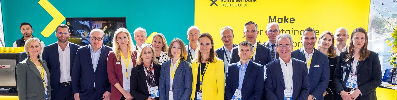Eine große Gruppe von Personen steht gemeinsam in einem Innenraum bei einer Firmenveranstaltung vor einem Hintergrund in Gelb und Türkis.