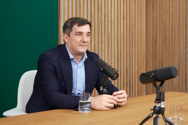 A man in a navy suit and light blue shirt sits at a wooden table with two microphones set up for a podcast or interview. The setting features a modern, minimalist design with vertical wooden slats and a green accent wall. Two glasses of water are visible on the table, and the overall mood is professional and conversational.