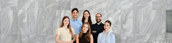 A group of six young adults stands together in front of a large gray marble wall. They are dressed in smart casual and business attire, suggesting a professional or corporate setting. The composition is centered on the people, with neutral stone textures filling the background. The overall mood is friendly, confident, and contemporary.