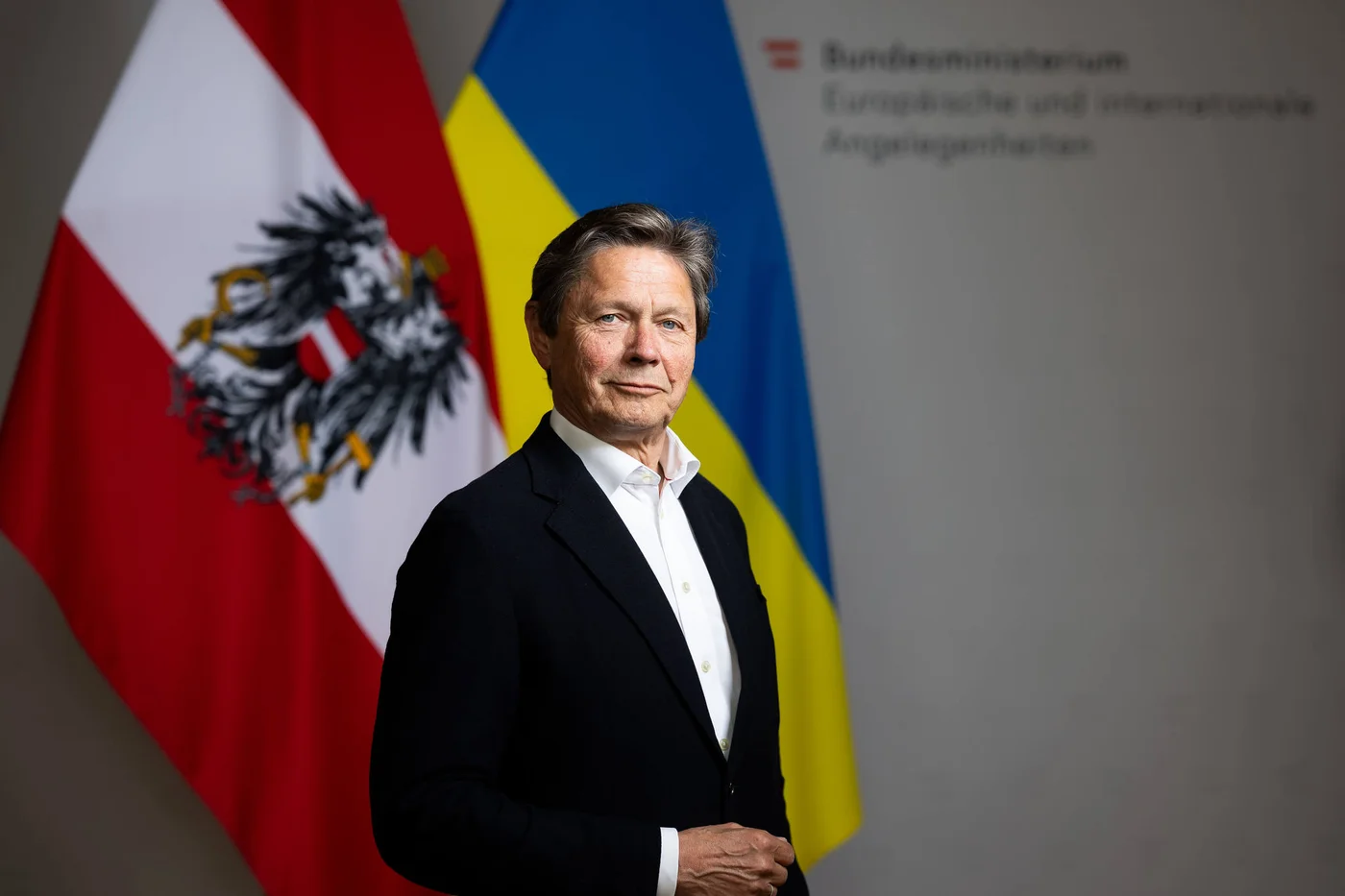 Eine Person in dunklem Anzug und weißem Hemd steht vor zwei großen Nationalflaggen, einer österreichischen und einer ukrainischen. Im Hintergrund ist teilweise ein Schriftzug mit dem Hinweis auf das österreichische Bundesministerium für Europa und Internationale Angelegenheiten zu sehen. Die Situation wirkt formell und dürfte mit Diplomatie oder internationalen Beziehungen in Verbindung stehen.
