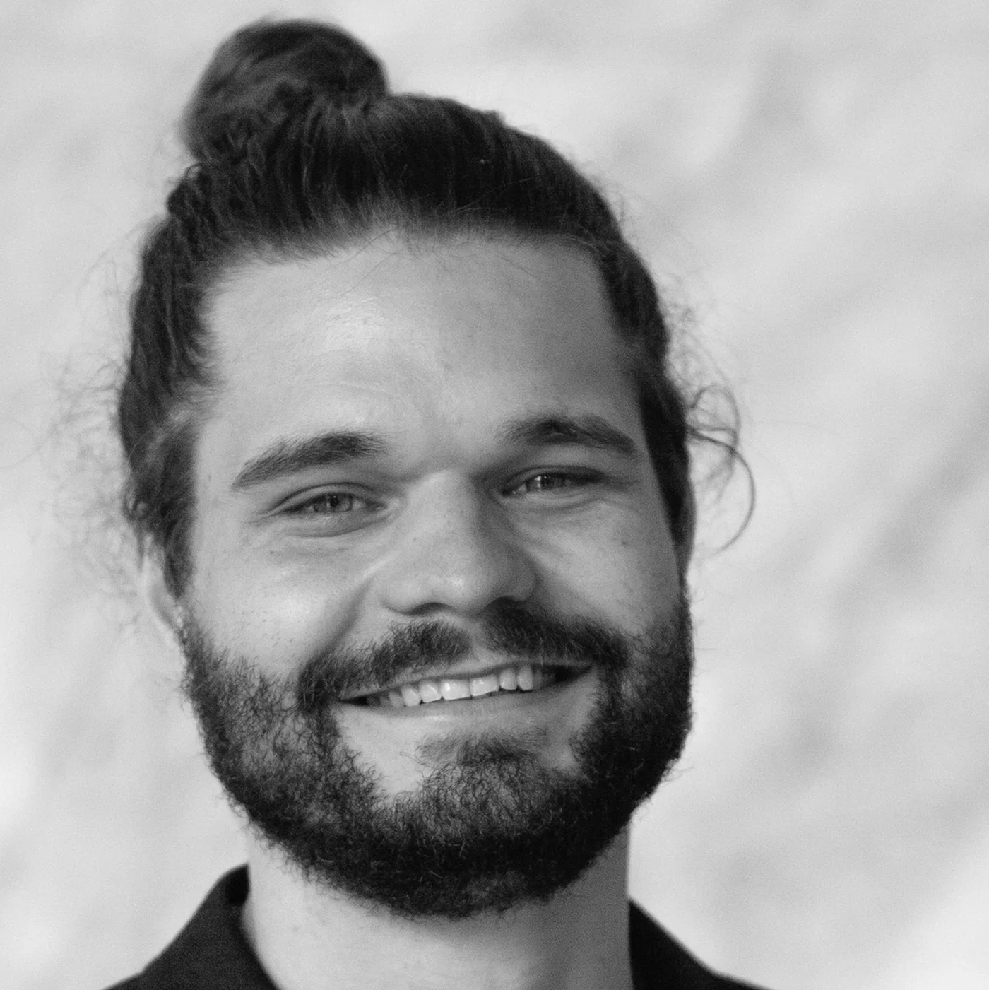 A black-and-white portrait of Moritz Welsner with long hair tied in a bun. The image features a neutral background with soft textures, creating a minimalist and timeless aesthetic. The subject is wearing a dark-colored shirt, adding contrast to the overall composition.