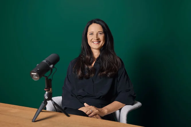 Marianne Heiß sitzt an einem Holztisch mit einem professionellen Mikrofon auf einem Stativ vor sich. Der Hintergrund ist eine einfarbig grüne Wand, die eine aufgeräumte und fokussierte Atmosphäre schafft. Das Mikrofon ist offenbar für Podcast- oder Aufnahmezwecke gedacht, und die Gesamtstimmung wirkt professionell und minimalistisch.