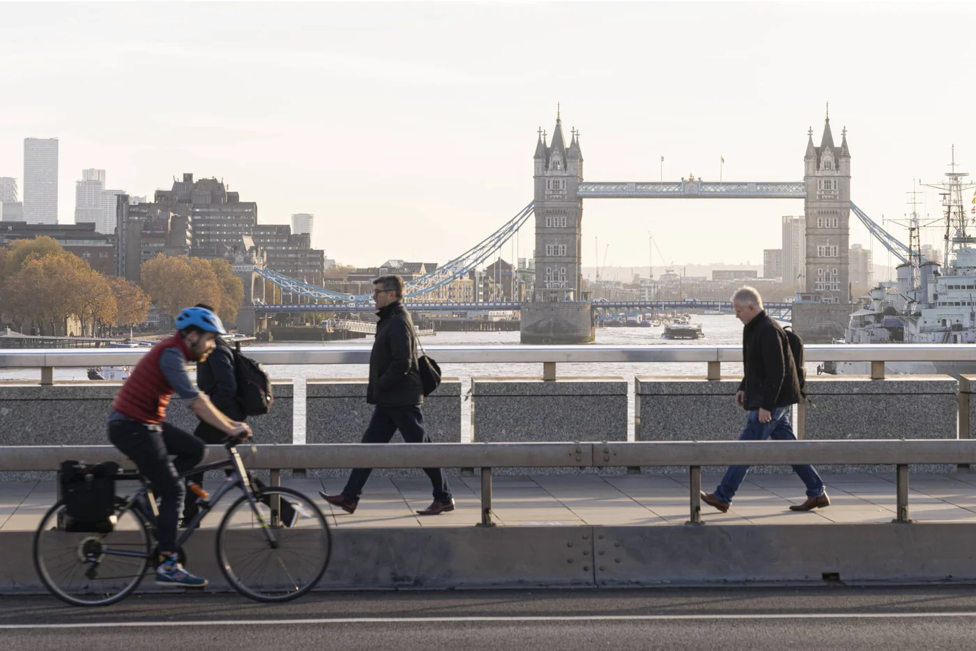 Eine Szene, die Fußgänger zeigt, die entlang eines Weges mit der Tower Bridge im Hintergrund spazieren. Ein Radfahrer fährt vorbei und schafft eine lebhafte städtische Atmosphäre, während die Sonne auf die Themse scheint.