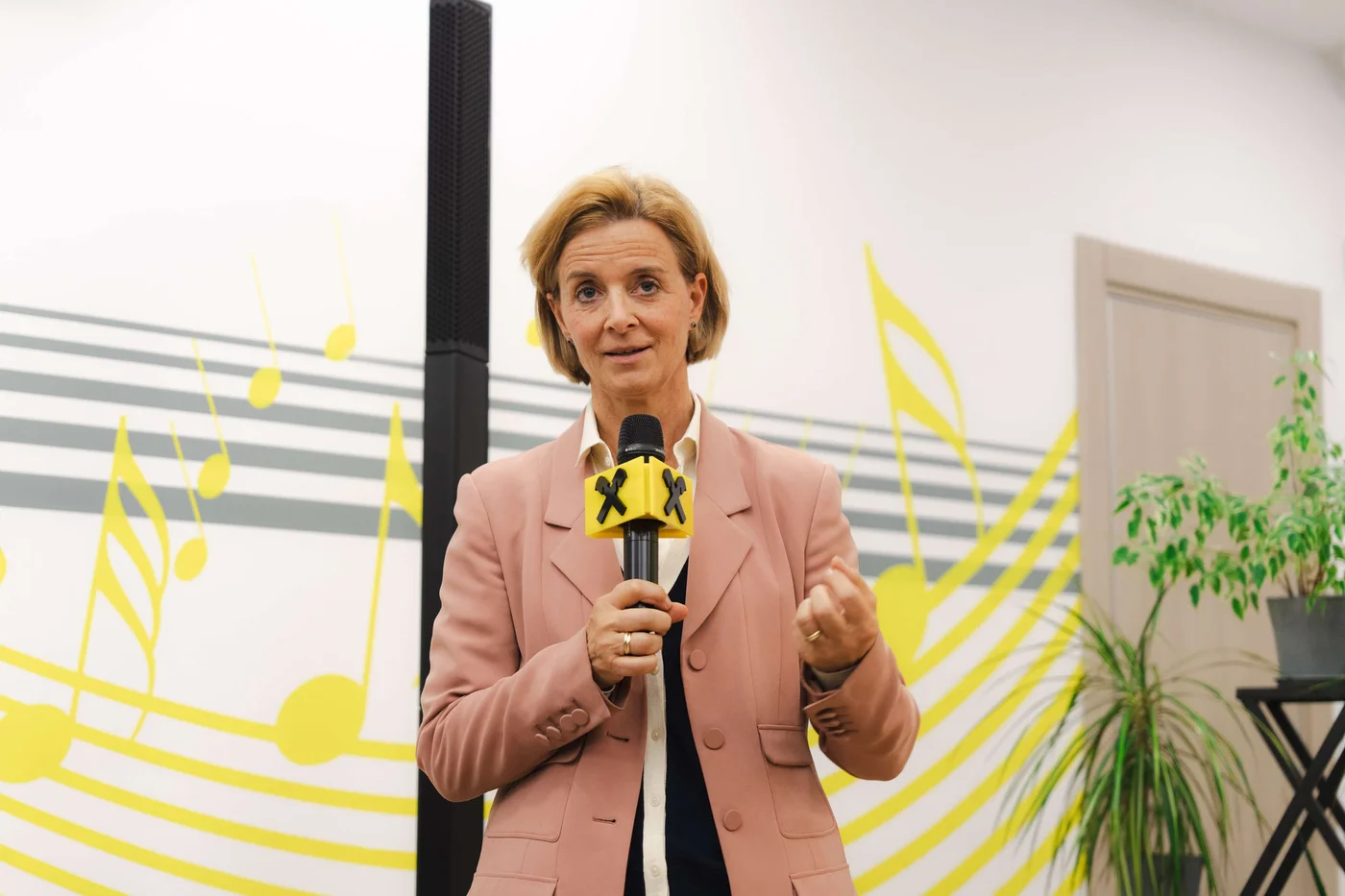 Eine Person in einem pinkfarbenen Blazer hält ein Mikrofon mit einem gelb-schwarzen „X“-Symbol und steht vor einer Wand, die mit Noten und Streifen verziert ist. Die Szenerie scheint in einem Innenraum zu sein; seitlich ist eine Topfpflanze und im Hintergrund eine Tür zu sehen. Die Stimmung ist professionell und lebendig, mit leuchtend gelben Akzenten und einer kreativen Atmosphäre. Es sind keine konkreten Zahlenwerte erkennbar.