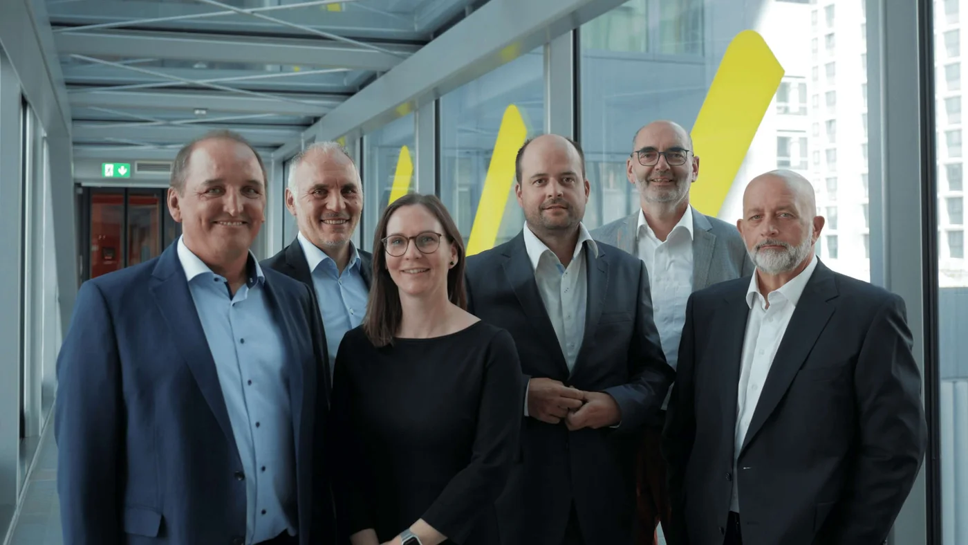 Sechs Geschäftsleute stehen in einem modernen Glaskorridor. Sie tragen formelle Businesskleidung, darunter Anzüge und ein dunkles Kleid, was auf ein Firmen- oder Führungsumfeld hindeutet. Große gelbe Formen sind an den Fenstern zu sehen, und im Hintergrund zeichnen sich Stadtgebäude ab. Die Stimmung ist professionell und selbstbewusst; die Gruppe posiert offenbar für ein Team- oder Führungsfoto.