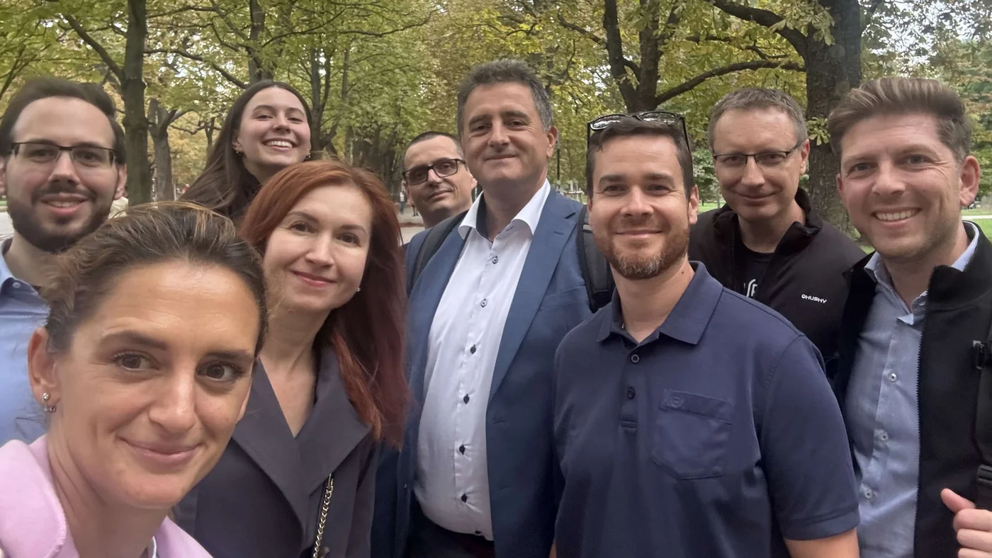 Eine Gruppe von Personen hat sich im Freien in einer parkähnlichen Anlage mit Bäumen und Grünflächen versammelt. Die Kleidung der Anwesenden ist eine Mischung aus legerer und formeller Kleidung, was auf ein privates oder berufliches Treffen hindeutet. Die Szene spielt am Tag, und natürliches Licht fällt durch die Bäume.