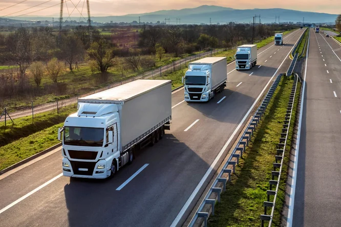 Ein Konvoi moderner Sattelschlepper fährt bei Sonnenuntergang eine mehrspurige Autobahn entlang und veranschaulicht so effiziente Gütertransporte und Logistikdienstleistungen über große Entfernungen.